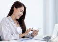 ノートパソコンの前でスマートフォンを操作する女性の画像